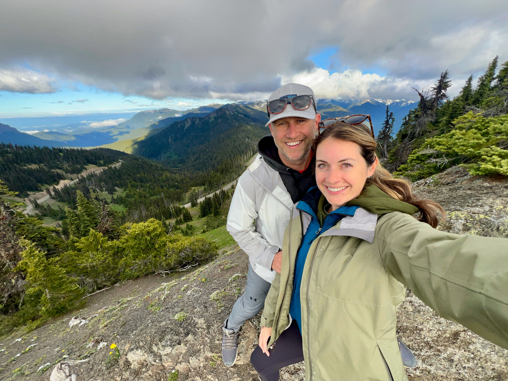 A couple taking a selfie with the rolling green hills and expansive view of the Olympic Mountains behind them at Hurricane Ridge.