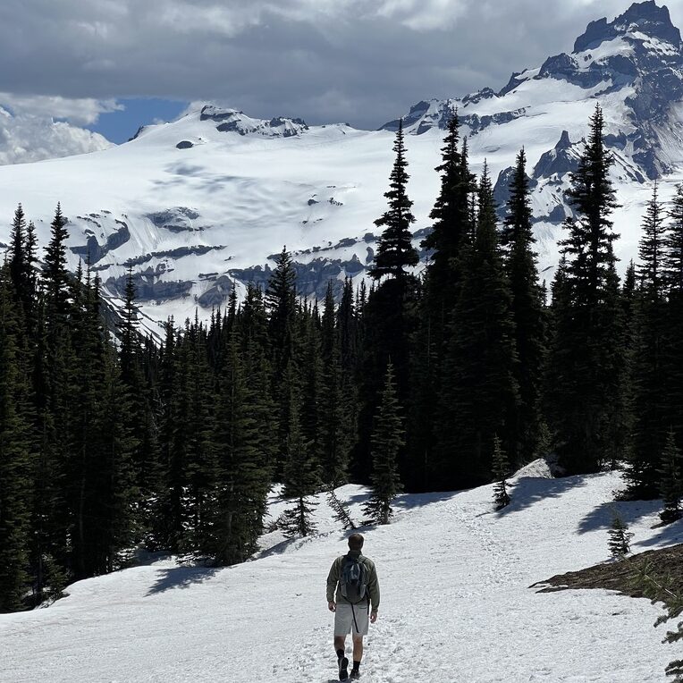 A hiker walking through a snow-covered trail with Mount Rainier’s snow-capped peaks in the background, framed by evergreen trees.