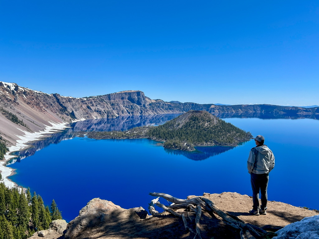 A hiker admiring the deep blue waters of Crater Lake with Wizard Island in the background, under a clear blue sky.