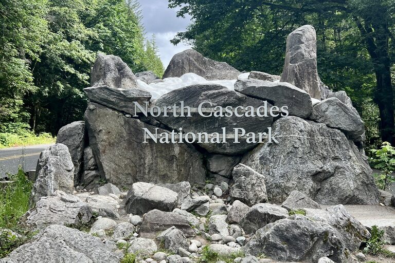 Large boulder with the text 'North Cascades National Park' engraved, surrounded by smaller rocks and lush green forest, with a cloudy sky in the background