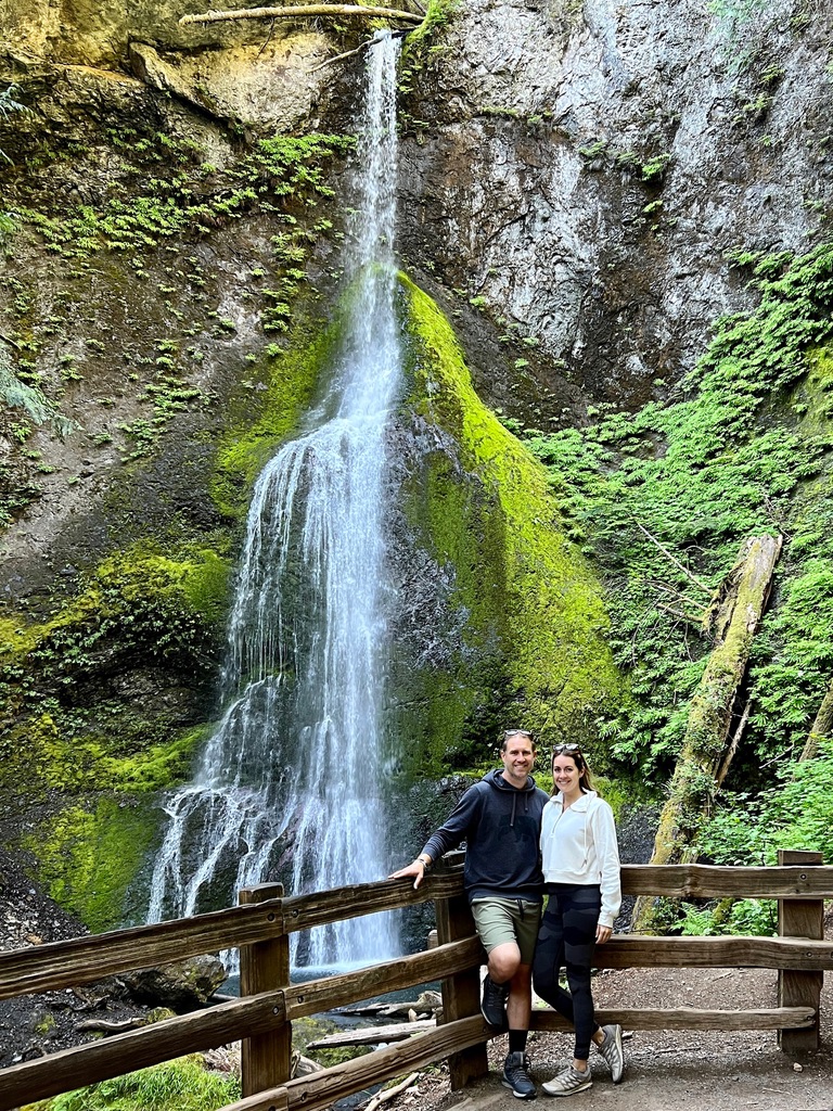 A couple posing on a wooden bridge in front of the cascading Marymere Falls in Olympic National Park surrounded by lush green moss and ferns.