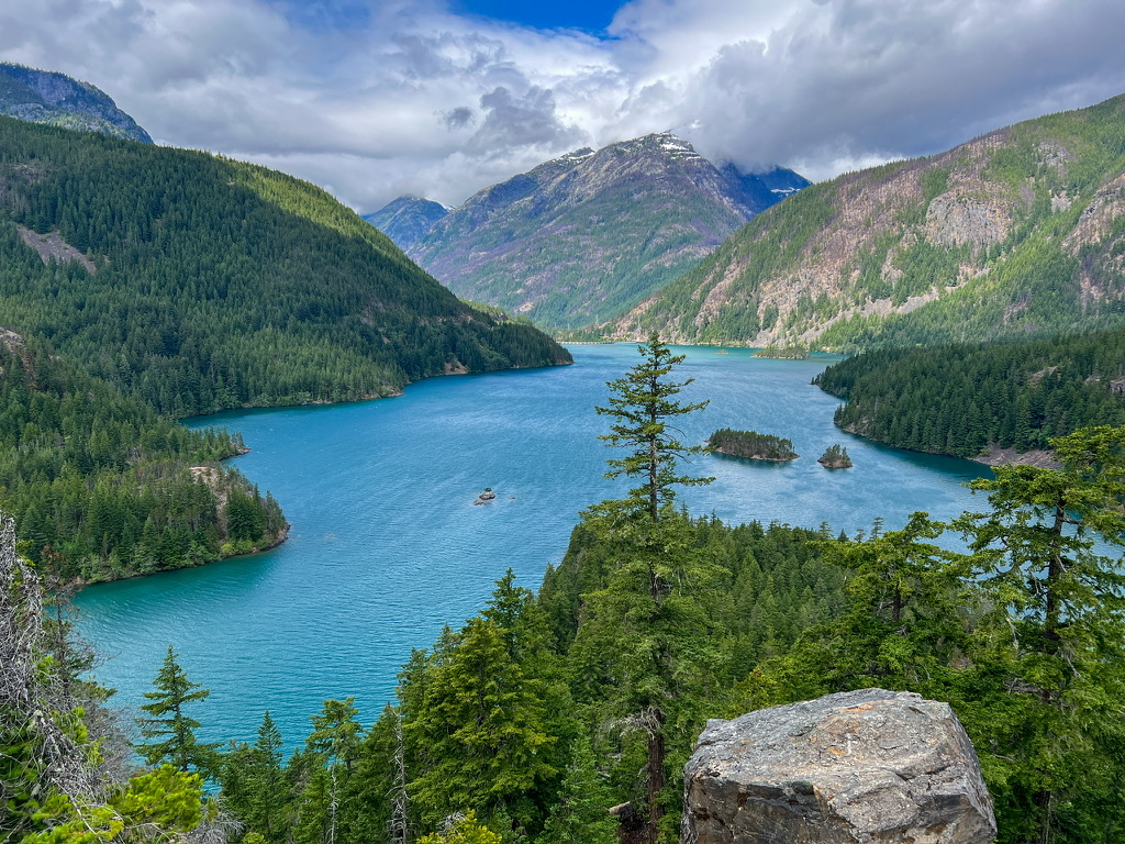 View of North Cascades National Park Diablo Lake's vibrant turquoise waters surrounded by dense evergreen forests and towering mountains, under a partly cloudy sky.