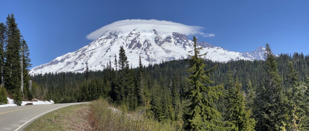 A scenic road winding through evergreen trees with a clear view of Mount Rainier, its peak draped in a lenticular cloud.