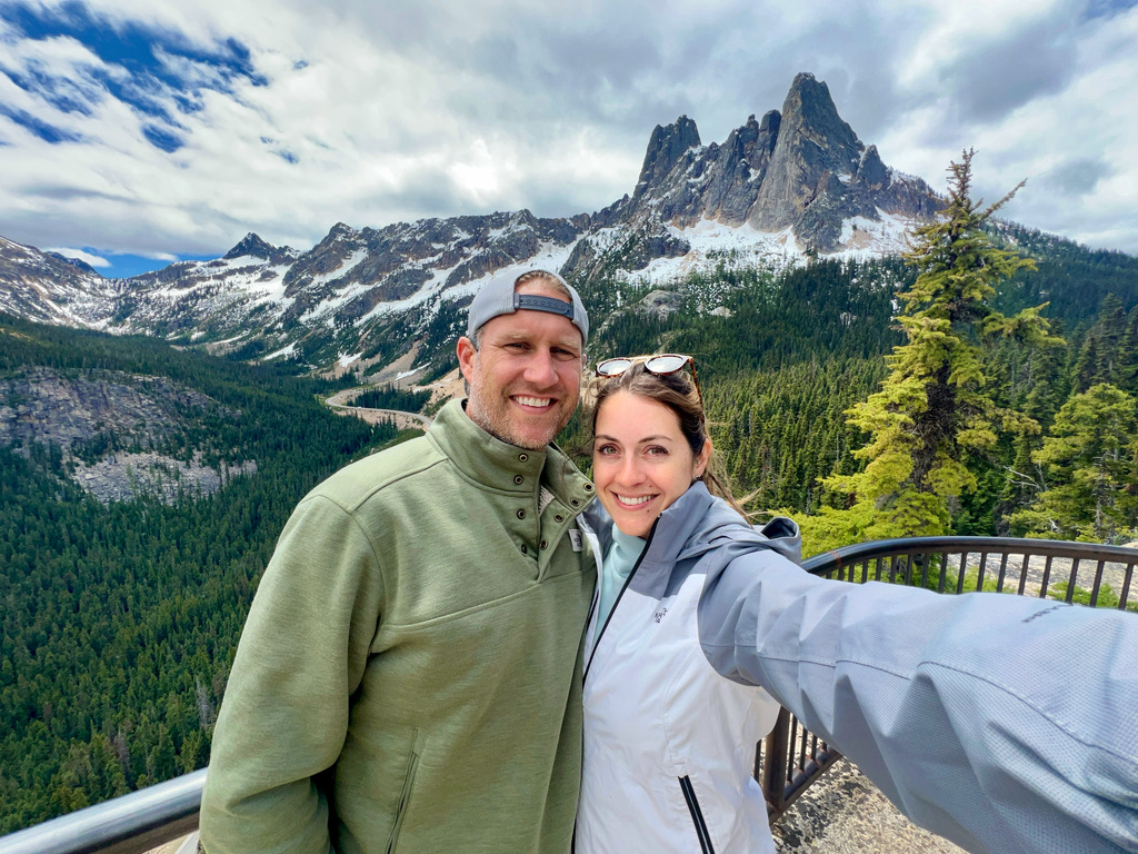 Couple smiling in front of the dramatic peaks of Liberty Bell Mountain at Washington Pass Overlook in North Cascades National Park, surrounded by lush green forests and a partly cloudy sky."