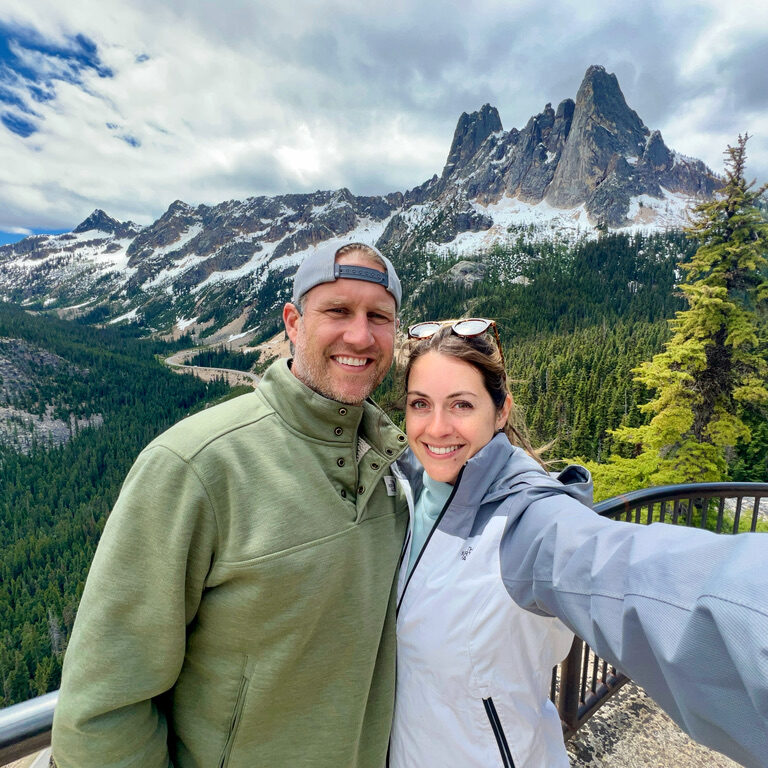 A couple poses for a selfie at the scenic overlook of Washington Pass in the North Cascades, surrounded by majestic, snow-capped peaks and dense forests. The man wears a green jacket and a backward cap, while the woman is dressed in a light gray jacket, both smiling against the backdrop of rugged mountains under a partly cloudy sky. The dramatic spires of Liberty Bell Mountain rise prominently in the distance, adding to the breathtaking alpine view.