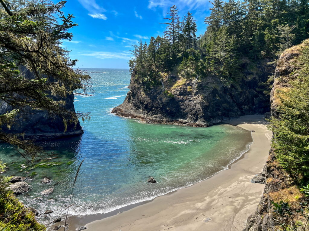 A secluded cove with crystal-clear turquoise water and sandy shores, surrounded by rugged cliffs and evergreen trees along the Samuel H. Boardman State Scenic Corridor on the Oregon Coast.