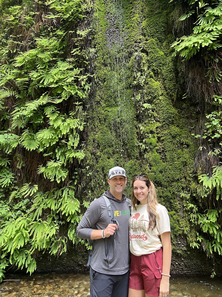 A couple standing in front of a moss-covered wall and small waterfall in Fern Canyon at Redwood National Park.