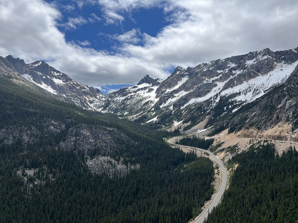 Scenic winding mountain road at Washington Pass, with jagged, snow-covered peaks and dense evergreen forests under a partly cloudy sky.