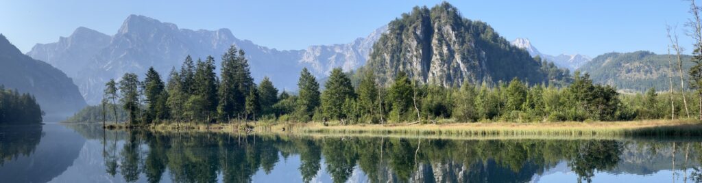 A panoramic view of Almsee lake in Austria, surrounded by lush greenery, towering rocky cliffs, and distant alpine peaks. The still, clear water reflects the vibrant trees and dramatic mountains, creating a serene and picturesque scene under a bright blue sky. The peaceful ambiance highlights the natural beauty of this alpine setting.