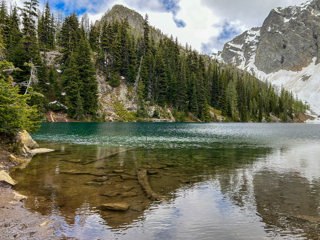 Crystal-clear alpine lake, Blue Lake in North Cascades National Park, reflecting the surrounding evergreen forest and rocky, snow-covered peaks on a calm, partly cloudy day.