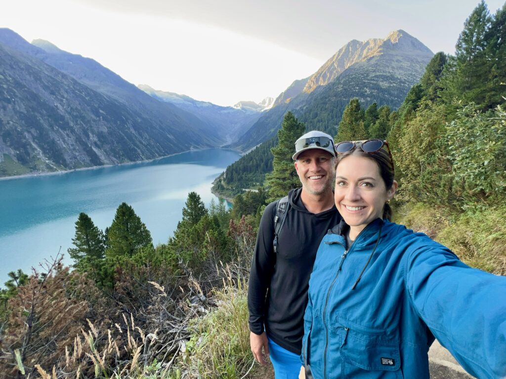 A smiling couple stands on the Olpererhütte trail in Austria with a stunning alpine lake and dramatic mountain peaks in the background. The lake's turquoise water is framed by lush green pine trees and steep rock slopes, creating a breathtaking scenic view. The couple is dressed in hiking gear, enjoying the natural beauty around them.