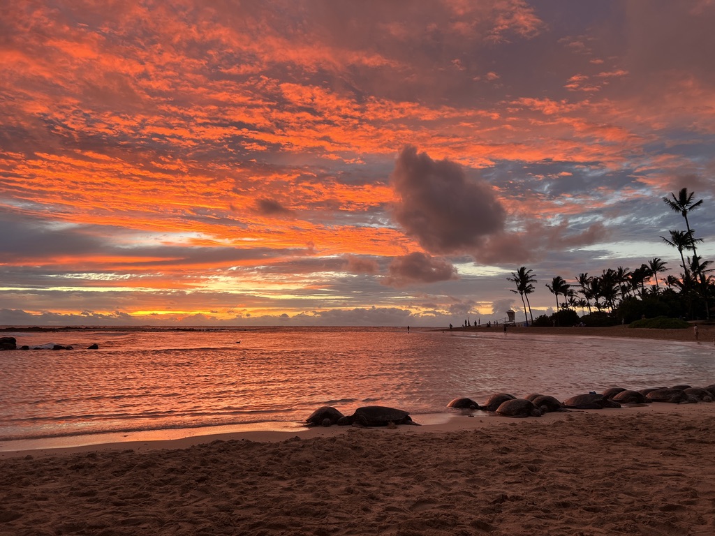 A breathtaking sunset at Poipu Beach in Kauai, Hawaii, with the sky ablaze in shades of orange, pink, and purple. Gentle waves reflect the vibrant colors, while palm trees and silhouettes of people add to the tranquil, tropical atmosphere. The sandy shoreline and scattered rocks frame this serene and stunning scene.