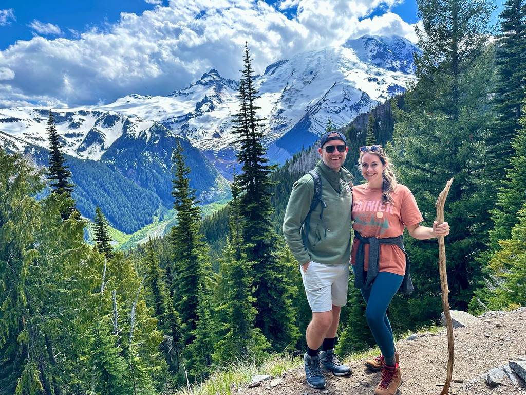 Two hikers posing with Mount Rainier in the background, its snowy peak towering above a lush green valley and evergreen trees.