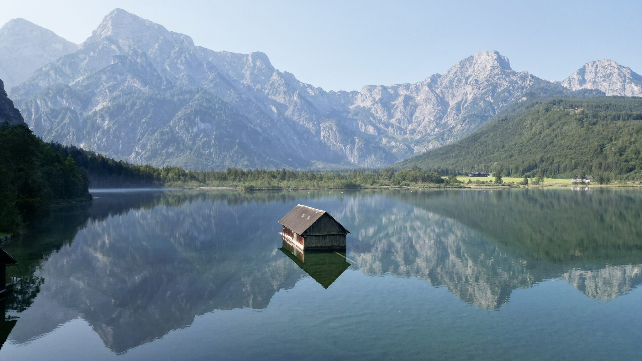 A serene view of Almsee lake in Austria, surrounded by towering mountains and lush green forests. The crystal-clear water reflects the rugged peaks and a quaint wooden boathouse, creating a tranquil and picturesque scene. The soft morning light enhances the peaceful ambiance of this alpine landscape.