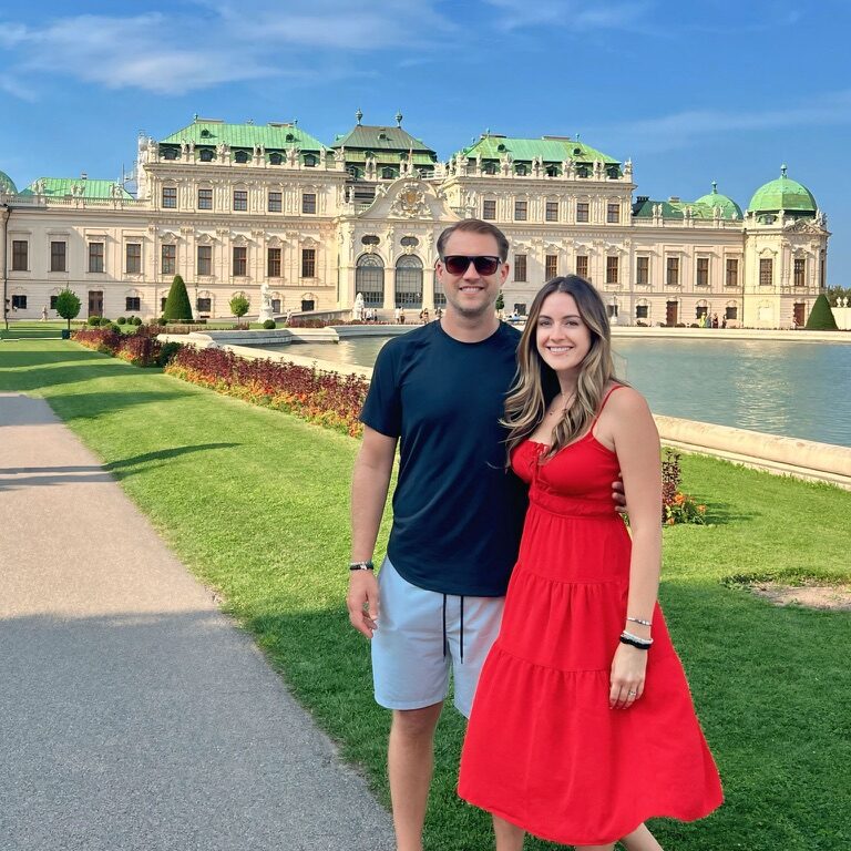 A couple poses in front of the grand Belvedere Palace in Vienna, Austria, with its ornate architecture and green domed roofs. The woman wears a bright red dress, and the man is dressed in casual summer attire. The lush gardens, manicured lawns, and a reflecting pond add to the elegant and vibrant setting on a sunny day.