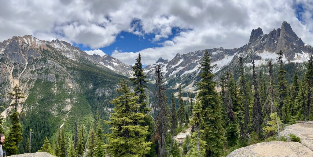 Panoramic view of Washington Pass in North Cascades National Park, featuring jagged snow-covered peaks, dense evergreen forests, and a winding mountain road under a bright sky with clouds