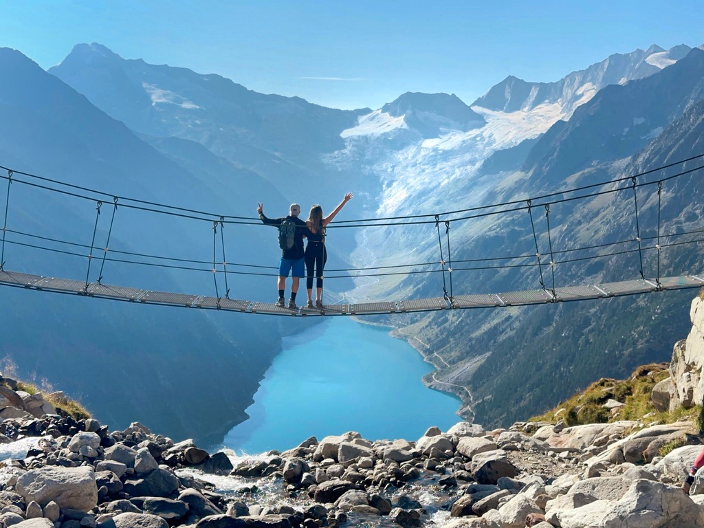 A couple stands on Olpererhütte suspension bridge (Panoramabrücke Olpererhütte) high above Lake Schlegeis, a vibrant blue alpine lake, with their arms raised in celebration. Surrounding them are dramatic mountain peaks and glaciers under a bright blue sky, creating a breathtaking and adventurous scene.