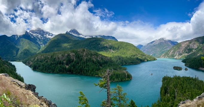 Panoramic view of Diablo Lake's vivid turquoise waters surrounded by lush green forests and rugged mountains under a bright blue sky with fluffy clouds.