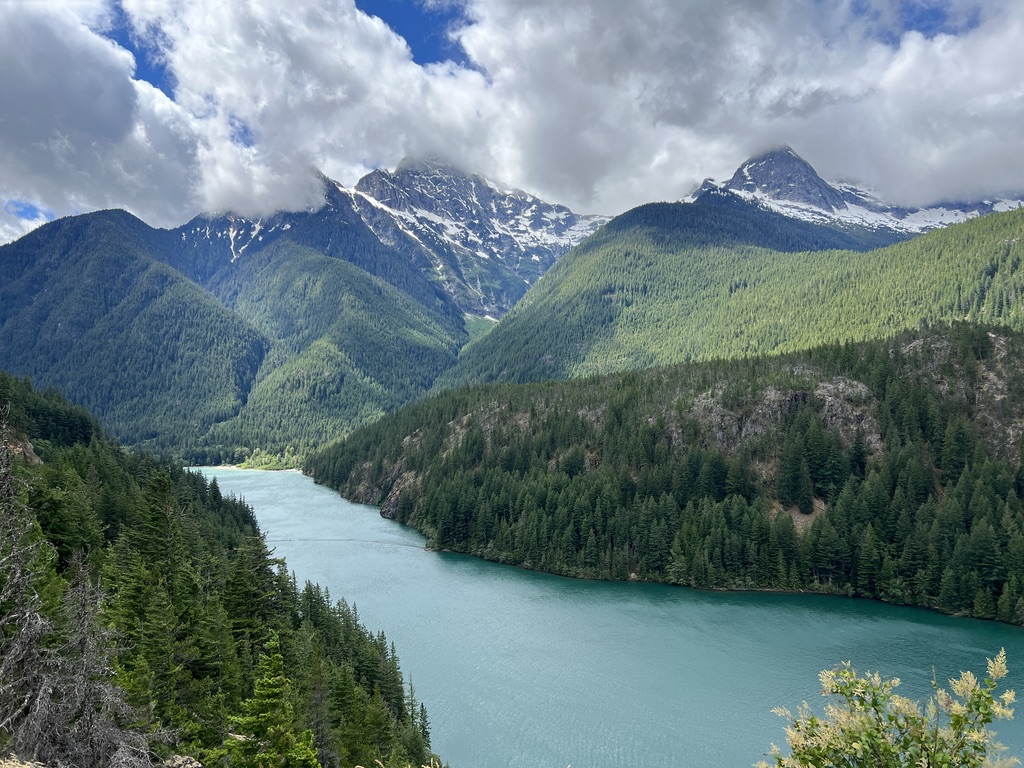 Scenic view of snow-capped mountains and lush green forests behind Diablo Lake, North Cascades National Park, under a dramatic cloudy sky.