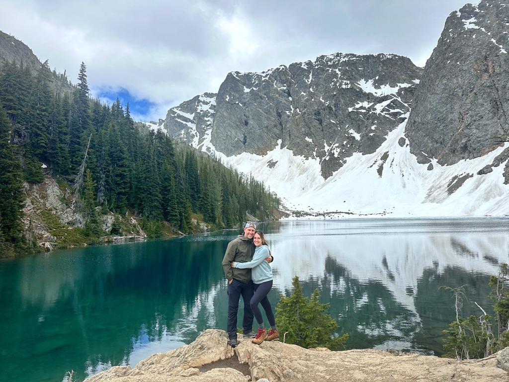 Couple standing on a rocky shoreline with Blue Lake behind them, surrounded by tall evergreen trees and towering, snow-covered mountains in North Cascades National Park