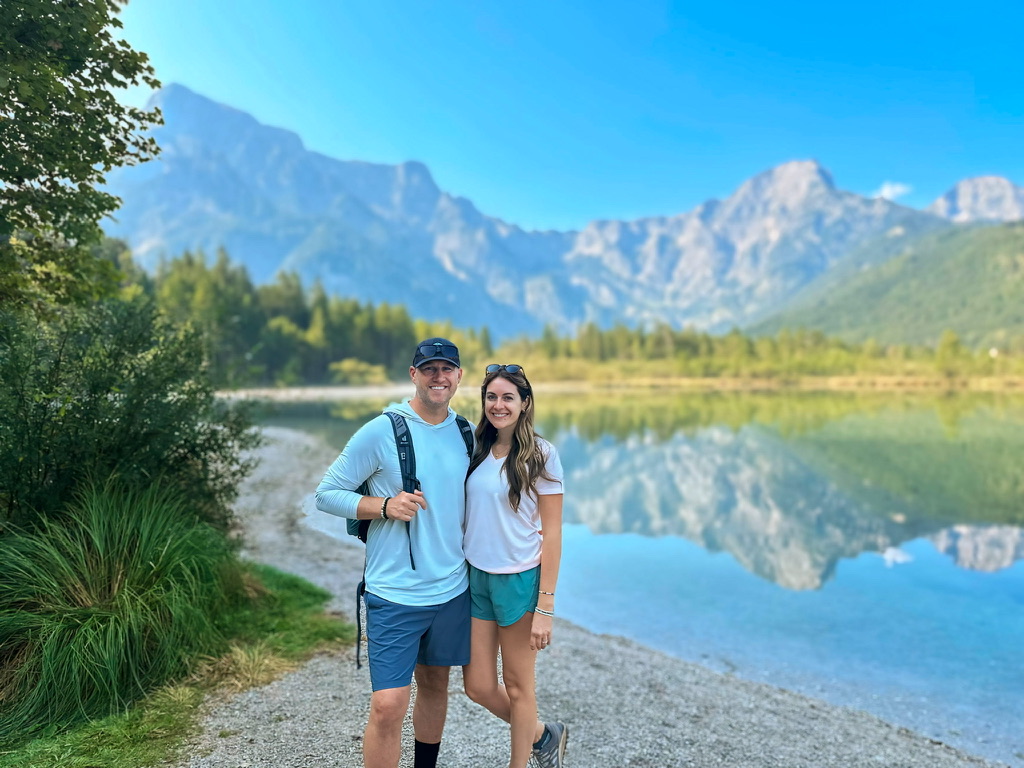 A smiling couple stands by the edge of Almsee Lake in Austria, surrounded by lush greenery and majestic mountain peaks. The crystal-clear water reflects the vibrant blue sky and the stunning alpine landscape, creating a serene and picturesque backdrop.